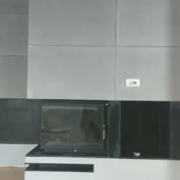 Nowoczesny kominek z obudową z szarych płyt, z czarnymi elementami wentylacyjnymi i wkładem kominkowym z przeszklonymi drzwiczkami, na jasnej drewnianej podłodze.