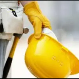 Robotnik w roboczych dżinsach, żółtej rękawicy i pasem narzędziowym trzyma żółty kask budowlany, widoczny młotek.