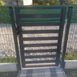 Nowoczesna, grafitowa furtka z poziomymi panelami, zamontowana przy ogrodzeniu z siatki, widok z zewnątrz na posesję z kostką brukową.