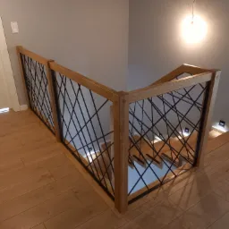 Nowoczesna balustrada z drewnianą ramą i czarnym, geometrycznym wypełnieniem, zamontowana przy schodach z drewnianymi stopniami i podświetleniem LED wzdłuż ściany.