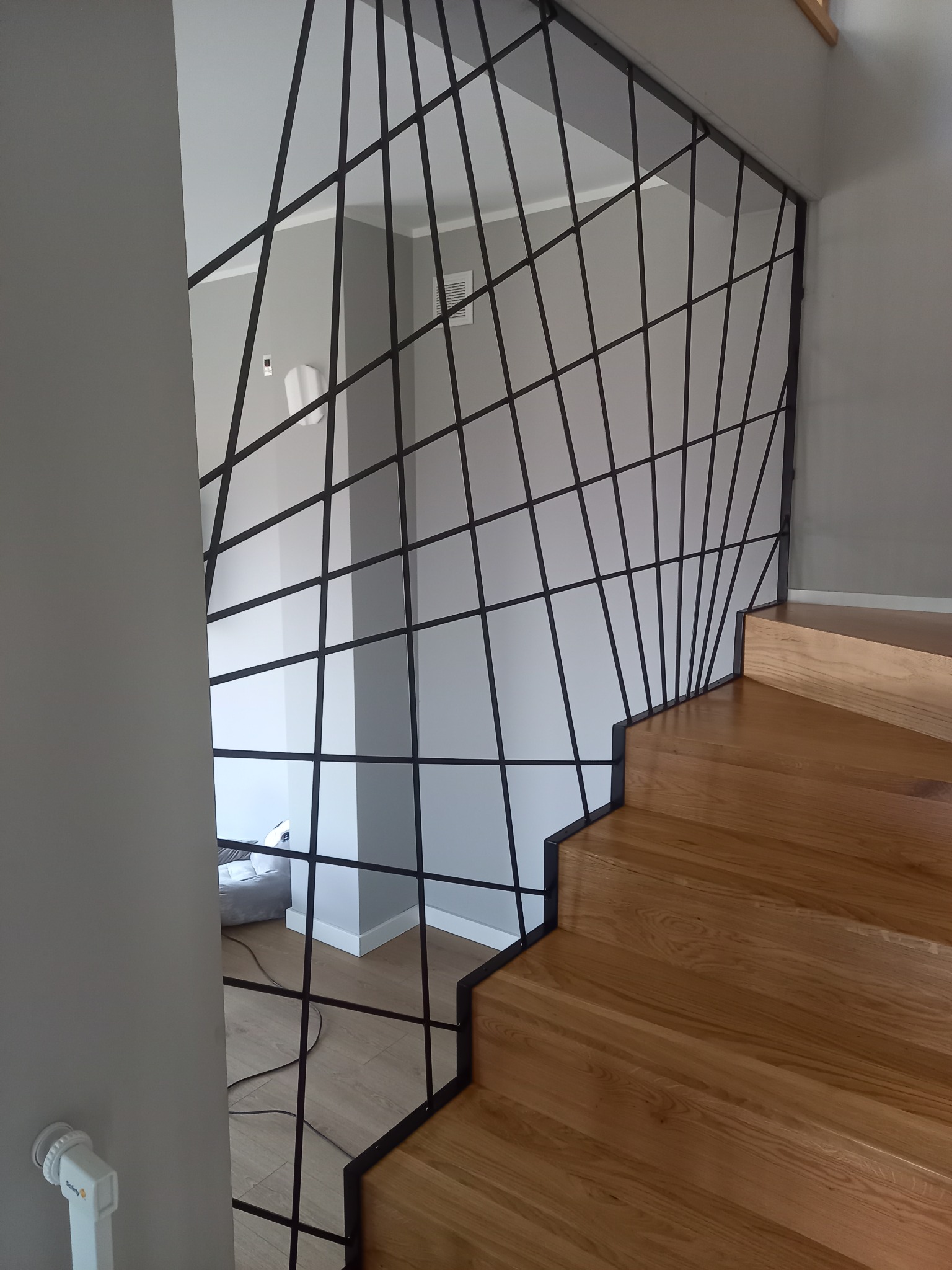 Nowoczesna balustrada ze stali o geometrycznym wzorze, zabezpieczająca drewniane schody w jasnym wnętrzu.