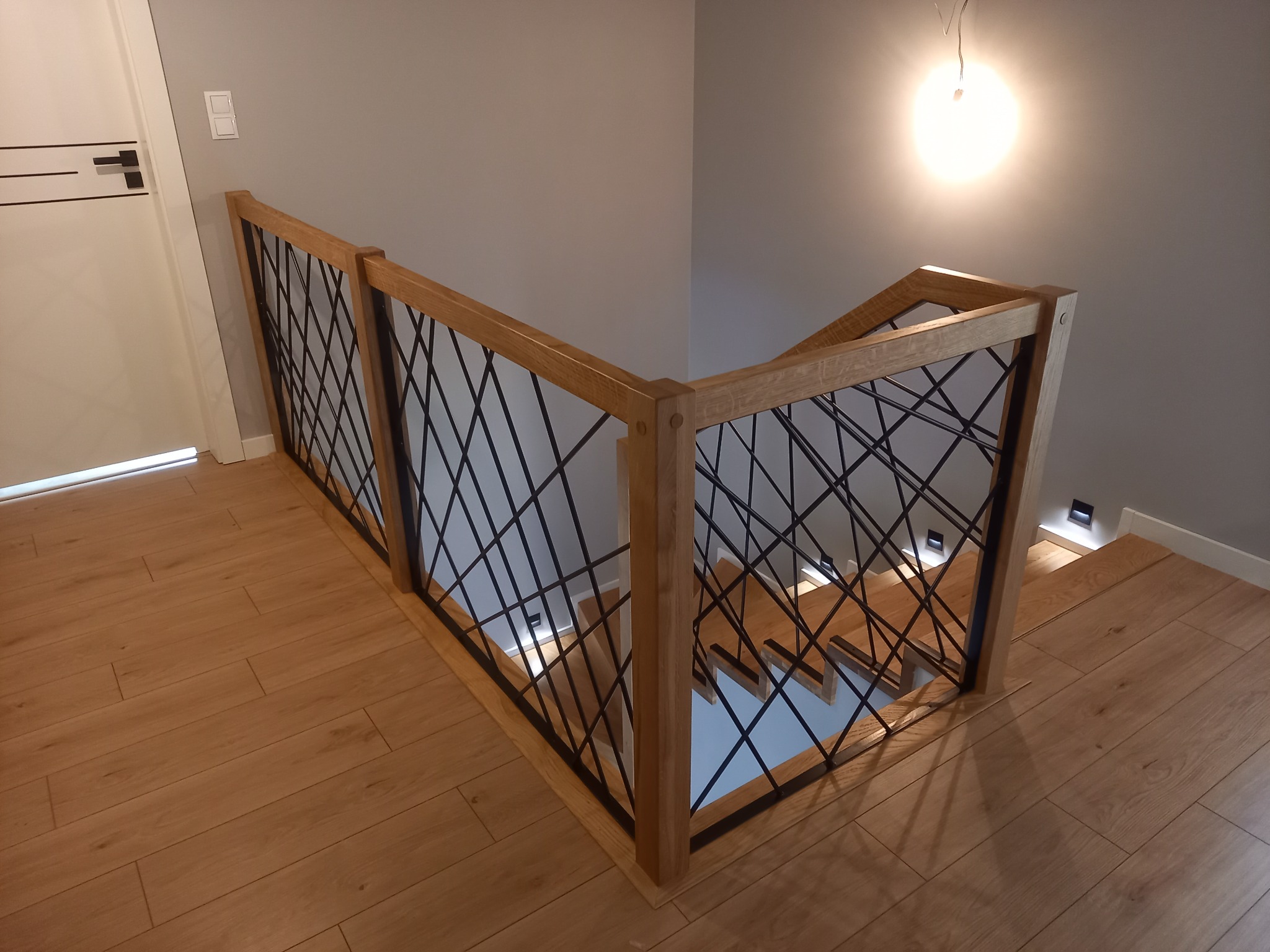 Nowoczesna balustrada z drewnianą ramą i czarnym, geometrycznym wypełnieniem, zamontowana przy schodach z drewnianymi stopniami i podświetleniem LED wzdłuż ściany.