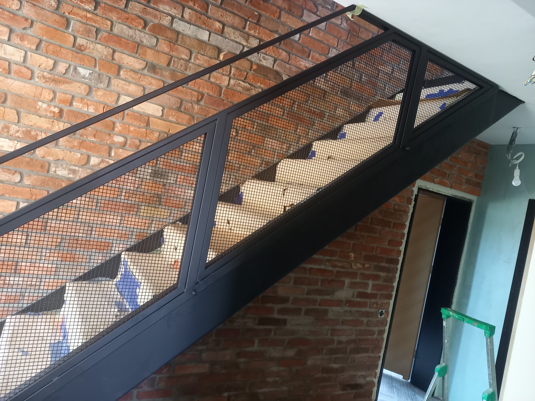 Nowoczesne schody z czarną metalową konstrukcją i siatkowym wypełnieniem, widok od dołu, na tle ściany z czerwonej cegły, zabezpieczone taśmą malarską na czas remontu.