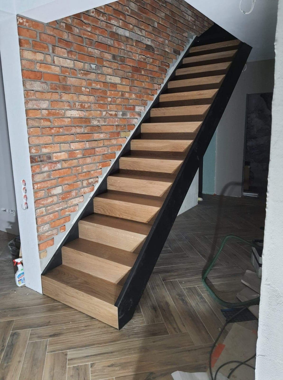 Nowoczesne schody drewniane z czarną metalową konstrukcją, przylegające do ściany z czerwonej cegły, na podłodze ułożonej w jodełkę.