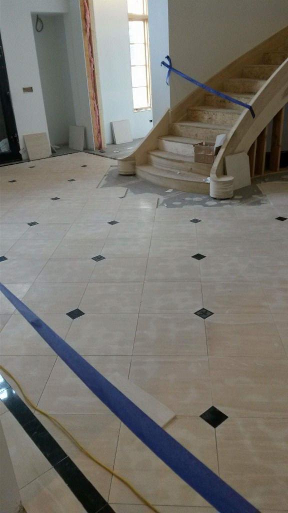 Podłoga z jasnych płytek z czarnymi wstawkami w trakcie układania, zabezpieczona niebieską taśmą malarską, widoczna drewniana konstrukcja schodów w tle.