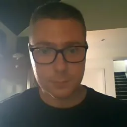 Mężczyzna w okularach na tle wnętrza domu z widocznymi schodami i elementami drewnianymi konstrukcji dachu.