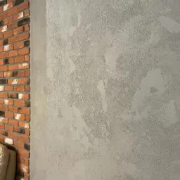 Szara ściana z efektem dekoracyjnym, obok fragment ściany z cegły, widoczny narożnik jasnej sofy.