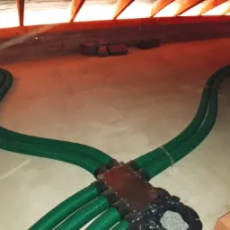 Zielone, karbowane rury wentylacyjne rozchodzące się w dwóch kierunkach w przestrzeni poddasza, z widoczną drewnianą konstrukcją dachu i cegłami w tle.