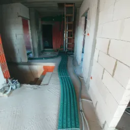 Wnętrze w trakcie budowy z widocznymi rzędami zielonych rur instalacyjnych prowadzonych po podłodze, białe ściany z betonu komórkowego, widoczne instalacje elektryczne w peszlach i drabina w tle.