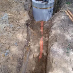 Betonowy zbiornik z włazem nad wykopem, w którym ułożono pomarańczowe rury, widoczne stopy osoby stojącej na zbiorniku, ziemia wokół wykopu.