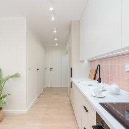 Holistic Studio Julia Ulikowska - Jasna kuchnia z różowymi płytkami na ścianie, białymi szafkami i blatem, czarną armaturą i płytą indukcyjną, oraz palmą w plecionym koszu w tle.