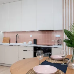Jasna kuchnia z beżowymi szafkami, różowymi płytkami i okrągłym drewnianym stołem z nakryciem na dwie osoby oraz dekoracyjną rośliną w szklanym wazonie.