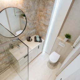 Holistic Studio Julia Ulikowska - Jasna łazienka z kabiną prysznicową ze szklaną ścianką, okrągłym lustrem, umywalką nablatową, białymi szafkami i toaletą w zabudowie, wykończona płytkami w odcieniach beżu i błękitu.