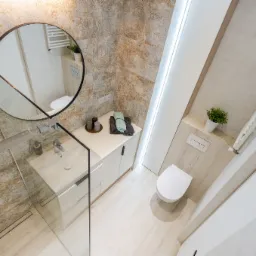 Jasna łazienka z kabiną prysznicową ze szklaną ścianką, okrągłym lustrem, umywalką nablatową, białymi szafkami i toaletą w zabudowie, wykończona płytkami w odcieniach beżu i błękitu.