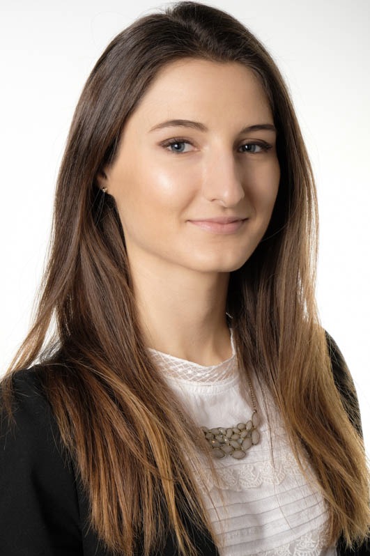 Portret młodej kobiety z długimi brązowymi włosami, ubrana w białą bluzkę z koronkowym wykończeniem i czarną marynarkę, na białym tle.