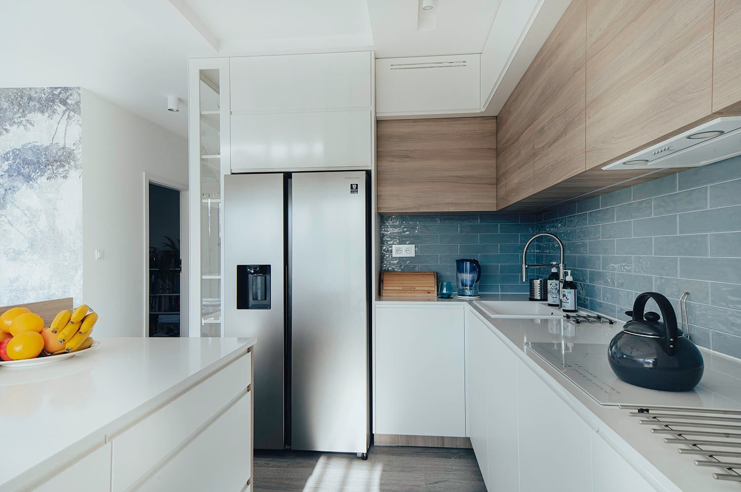 Jasna kuchnia z białymi szafkami, drewnianymi akcentami i niebieskimi płytkami na ścianie, wyposażona w dużą, srebrną lodówkę i czajnik na blacie.