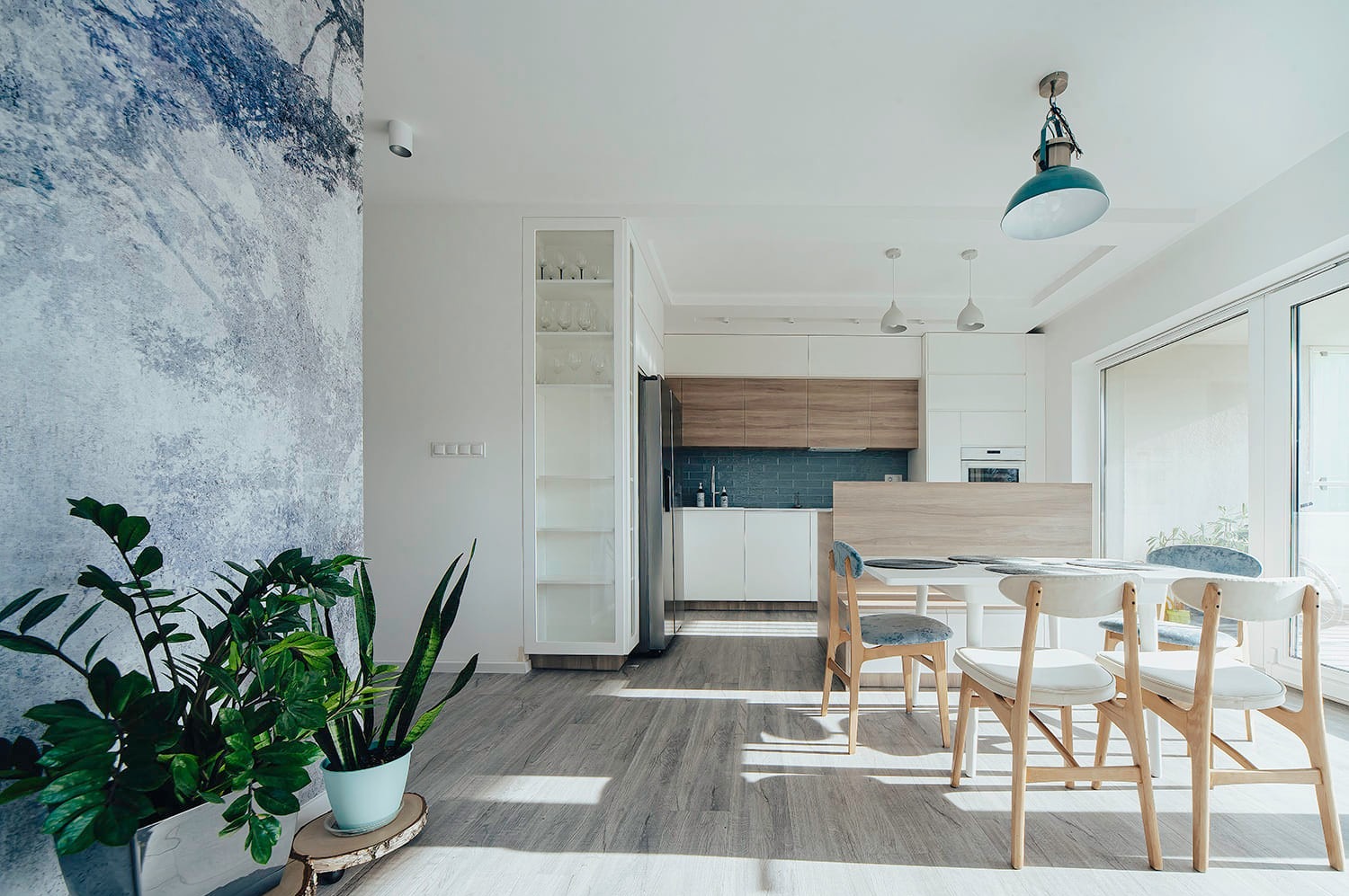 Jasne wnętrze mieszkania z drewnianą podłogą, ścianą z motywem drzewa, stołem z krzesłami i widokiem na kuchnię z niebieskimi płytkami.