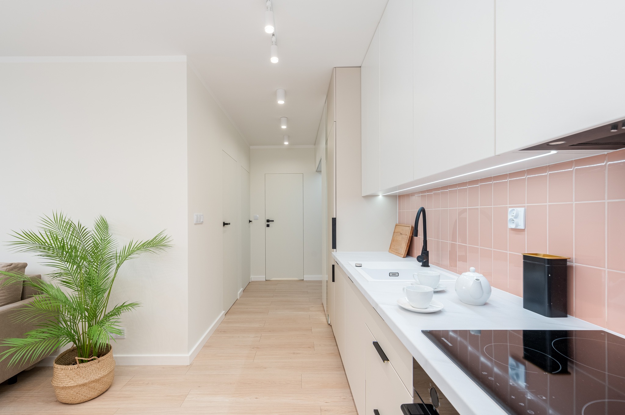 Jasna kuchnia z różowymi płytkami na ścianie, białymi szafkami i blatem, czarną armaturą i płytą indukcyjną, oraz palmą w plecionym koszu w tle.