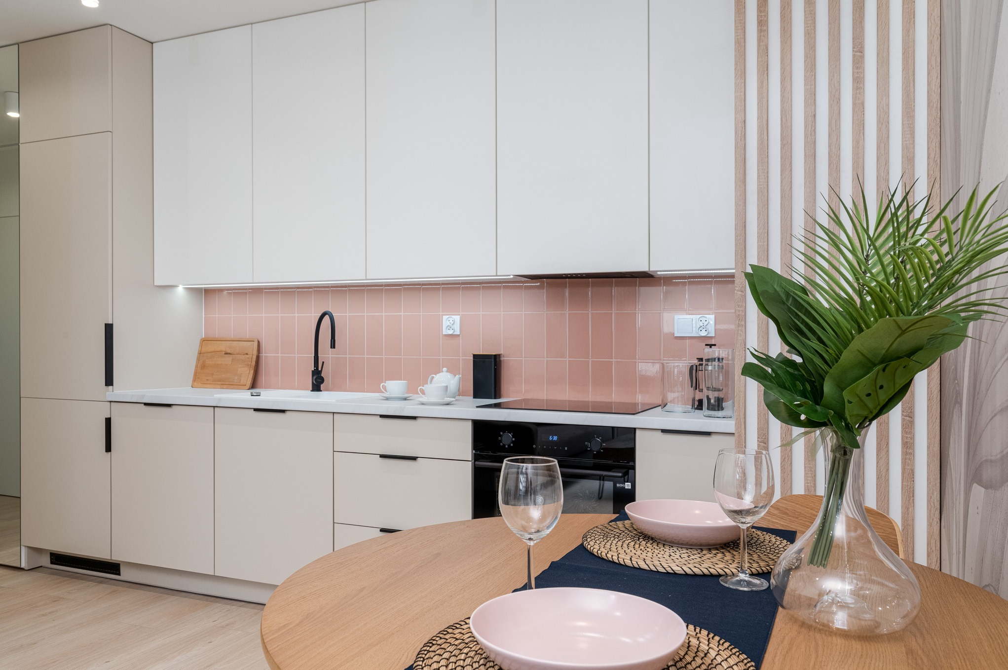 Jasna kuchnia z beżowymi szafkami, różowymi płytkami i okrągłym drewnianym stołem z nakryciem na dwie osoby oraz dekoracyjną rośliną w szklanym wazonie.