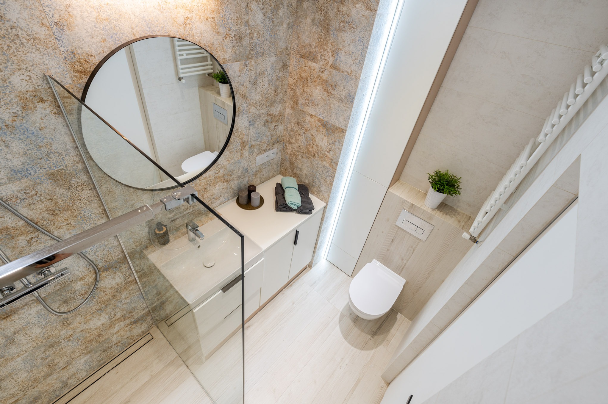 Jasna łazienka z kabiną prysznicową ze szklaną ścianką, okrągłym lustrem, umywalką nablatową, białymi szafkami i toaletą w zabudowie, wykończona płytkami w odcieniach beżu i błękitu.