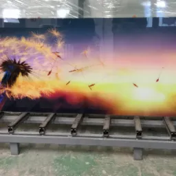 Duży, prostokątny panel ze szkła z nadrukiem przedstawiającym dmuchawca na tle zachodzącego słońca, umieszczony na metalowym stojaku.