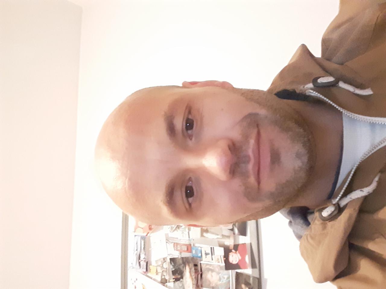 Portret mężczyzny z krótką brodą w brązowej kurtce na tle białej ściany z dekoracjami. Zdjęcie obrócone w prawo.