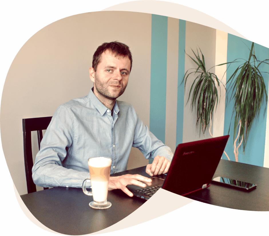 Mężczyzna w błękitnej koszuli siedzi przy biurku z laptopem, obok stoi latte macchiato i leży smartfon, w tle ściana w błękitne pasy i roślina doniczkowa.