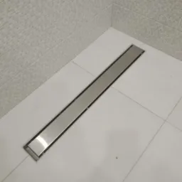 Dyskretny, liniowy odpływ prysznicowy ze stali nierdzewnej, wkomponowany w białe płytki podłogowe i mozaikę ścienną w narożniku łazienki.