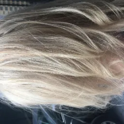 Rozjaśnione, proste włosy w odcieniach blondu, widoczne z góry, z delikatnym prześwitem skóry głowy i fragmentem niebieskiej koszuli w paski.