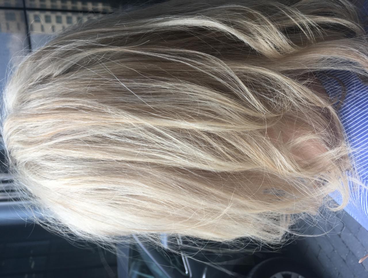 Rozjaśnione, proste włosy w odcieniach blondu, widoczne z góry, z delikatnym prześwitem skóry głowy i fragmentem niebieskiej koszuli w paski.