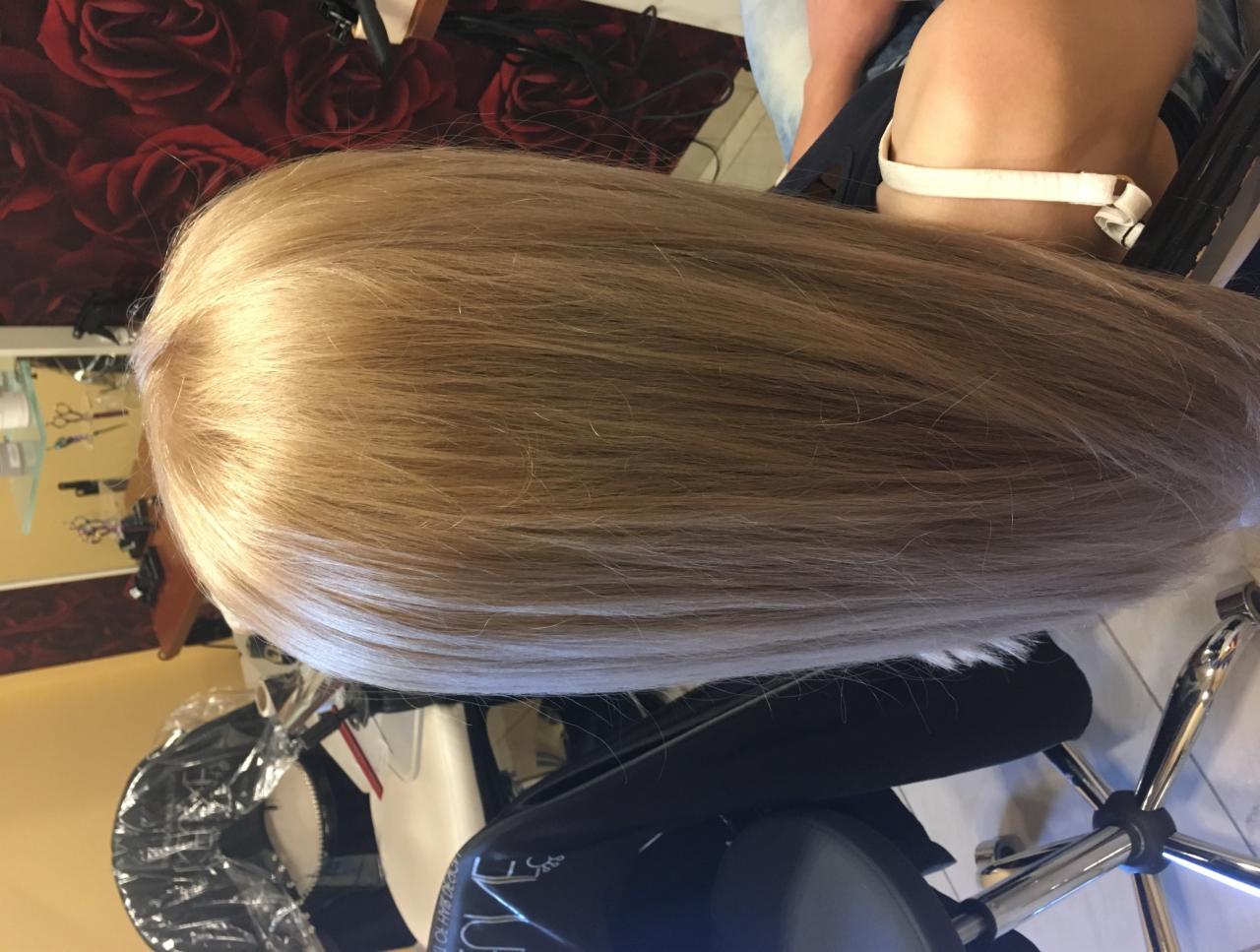 Widok z góry na blond włosy klientki w salonie, założony kołnierz ochronny, w tle lustro i narzędzia.