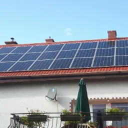 Panele słoneczne na dachu domu z czerwoną dachówką, widoczne anteny satelitarne i balkon z kwiatami.