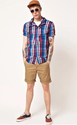 Mężczyzna w kraciastej koszuli z krótkim rękawem, beżowych szortach i brązowej czapce bejsbolówce, prezentujący letnią stylizację z tatuażami na ramionach i okularami przeciwsłonecznymi.