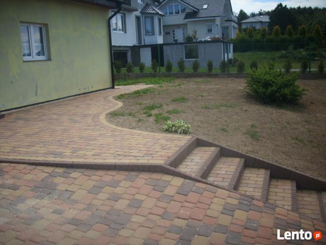 Ułożona, wielobarwna kostka brukowa prowadząca od ściany domu do schodów na trawnik. Widoczny zakrzywiony brzeg kostki i trzy stopnie schodów wykonane z tego samego materiału.