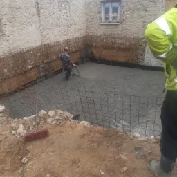 Wypełniony betonem wykop pod fundamenty zbrojony siatką, widoczny pracownik zacierający powierzchnię betonu, fragment ściany budynku w tle, narzędzia na ziemi.