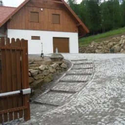 Ułożona kostka brukowa tworząca podjazd do domu z bramą wjazdową. Widoczne schody z kostki i kamienne umocnienie skarpy. Całość w otoczeniu zieleni.