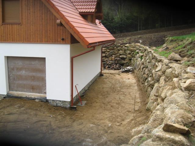 Budowa kamiennego muru oporowego przy budynku z garażem. Widoczne przygotowane podłoże i materiały budowlane. Ujęcie z ziemi, wczesna faza realizacji.