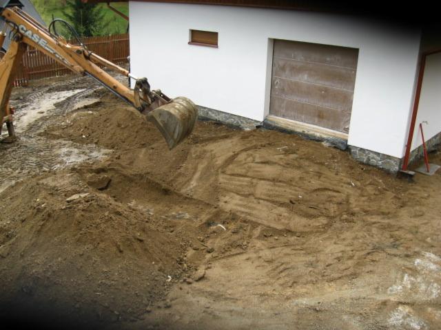 Żółta koparka CASE wyrównuje teren przy budynku z brązowymi drzwiami garażowymi, przygotowując podłoże pod dalsze prace budowlane. Widoczny świeżo usypany grunt.