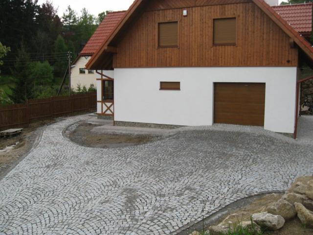 Ułożony z szarej kostki granitowej podjazd do domu z brązową bramą garażową. Kostka ułożona w łukowaty wzór. Widoczne fragmenty ogrodu i drewniany płot.