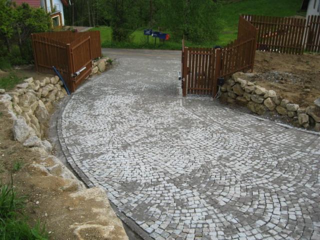 Podjazd z jasnej kostki granitowej ułożonej w łuk, z drewnianą bramą w tle i kamiennym murkiem po lewej stronie. Widok na posesję z bliska.