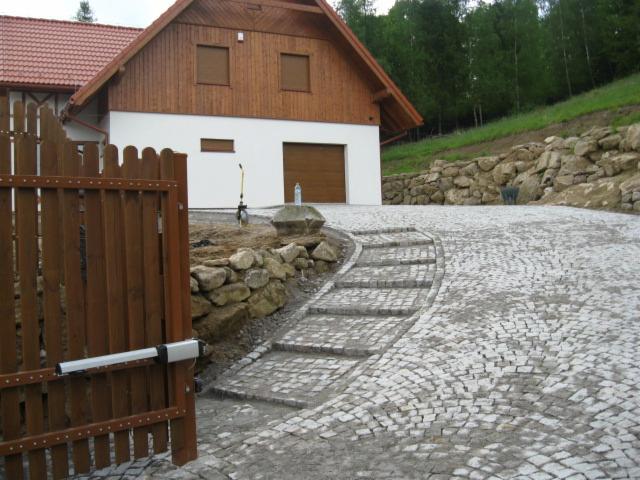 Ułożona kostka brukowa tworząca podjazd do domu z bramą wjazdową. Widoczne schody z kostki i kamienne umocnienie skarpy. Całość w otoczeniu zieleni.