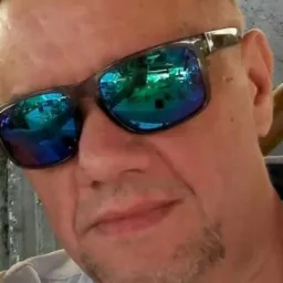 Mężczyzna w okularach przeciwsłonecznych z odbiciem wnętrza budynku. Ujęcie od dołu, widoczna część jasnej koszuli i fragment drewnianej konstrukcji.