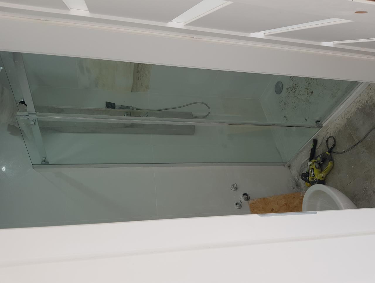 Montaż szklanej kabiny prysznicowej z przesuwnymi drzwiami; widoczna biała glazura, częściowo wykończona podłoga, narzędzia Ryobi i fragment umywalki.