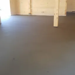 Świeżo wylana, gładka posadzka w surowym drewnianym domu, widoczne drewniane ściany i filary konstrukcyjne, przygotowana do dalszych prac wykończeniowych.