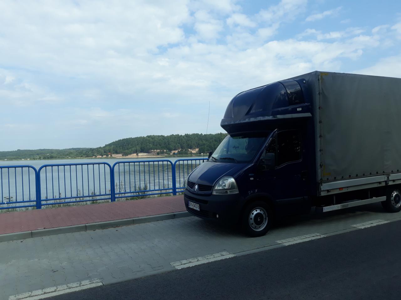 Niebieski bus z plandeką na tle jeziora i błękitnego nieba. Pojazd zaparkowany obok barierki, widoczny numer rejestracyjny. Transport towarów na trasach.