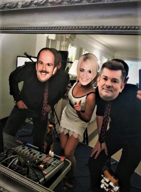 Odbicie w lustrze przedstawia troje osób z nałożonymi maskami na twarzach, w tle widoczny sprzęt muzyczny.