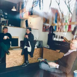 Uczestnicy kursu marketingu w Tychach podczas przerwy na kawę. Widoczny mężczyzna z laptopem i dwie kobiety z kubkami siedzące na schodach. W tle regał z książkami i dekoracje.