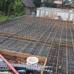 Zbrojenie płyty fundamentowej z widocznymi narzędziami i pracownikiem w tle, pod budowę domu jednorodzinnego.