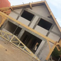 Surowy stan domu z bloczków, z widoczną drewnianą konstrukcją dachu i niezadaszonymi oknami, na tle błękitnego nieba.