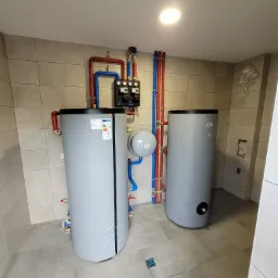 Dwa szare zbiorniki ciepłej wody użytkowej z podłączonymi rurami (czerwone i niebieskie) w kotłowni wykończonej płytkami ceramicznymi. Widoczne zawory i manometry.